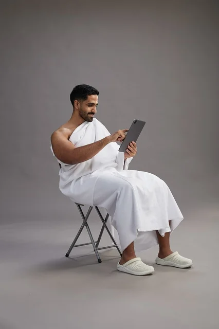 A portrait of a Saudi pilgrim in Ihram clothing holding a tablet and sitting on a chair, preparing to perform the rituals of Hajj and Umrah, with a gray background. A portrait of a Saudi pilgrim in Ihram clothing holding a tablet and sitting on a chair, preparing to perform the rituals of Hajj and Umrah, with a gray background.