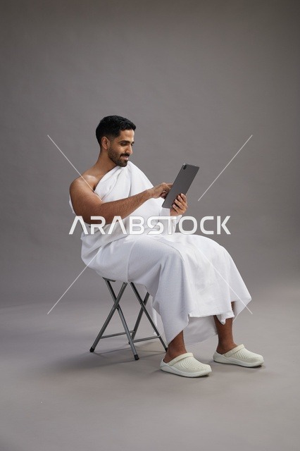 بورتريه لحاج سعودي بملابس الإحرام يحمل تابلت ويجلس على كرسي, الاستعداد لاداء مناسك الحج والعمره, خلفيه رماديه