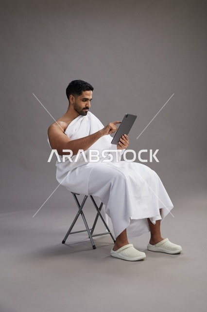 بورتريه لحاج سعودي بملابس الإحرام يحمل تابلت ويجلس على كرسي, الاستعداد لاداء مناسك الحج والعمره, خلفيه رماديه