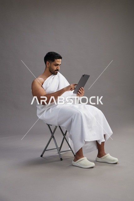 A portrait of a Saudi pilgrim in Ihram clothing holding a tablet and sitting on a chair, preparing to perform the rituals of Hajj and Umrah, with a gray background.