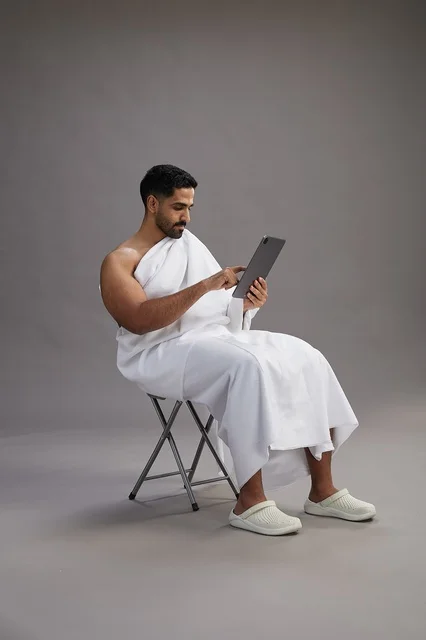 A portrait of a Saudi pilgrim in Ihram clothing holding a tablet and sitting on a chair, preparing to perform the rituals of Hajj and Umrah, with a gray background.
