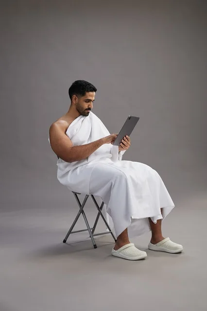 A portrait of a Saudi pilgrim in Ihram clothing holding a tablet and sitting on a chair, preparing to perform the rituals of Hajj and Umrah, with a gray background.