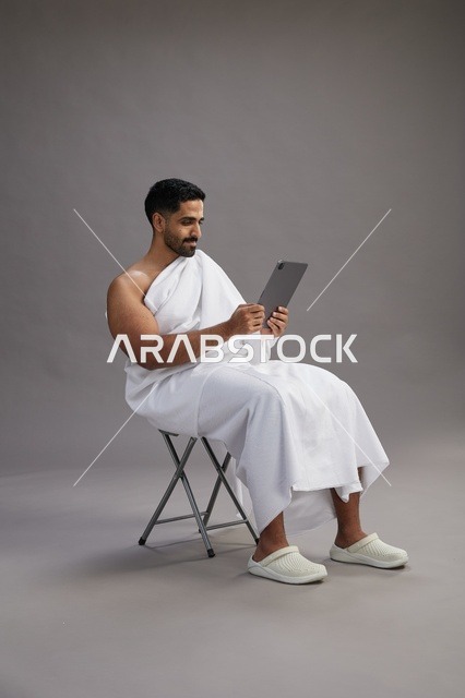 بورتريه لحاج سعودي بملابس الإحرام يحمل تابلت ويجلس على كرسي, الاستعداد لاداء مناسك الحج والعمره, خلفيه رماديه
