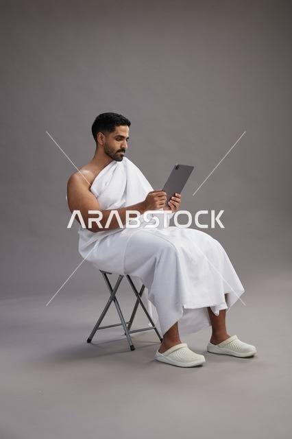 بورتريه لحاج سعودي بملابس الإحرام يحمل تابلت ويجلس على كرسي, الاستعداد لاداء مناسك الحج والعمره, خلفيه رماديه