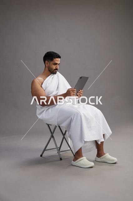A portrait of a Saudi pilgrim in Ihram clothing holding a tablet and sitting on a chair, preparing to perform the rituals of Hajj and Umrah, with a gray background.