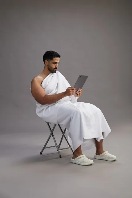 A portrait of a Saudi pilgrim in Ihram clothing holding a tablet and sitting on a chair, preparing to perform the rituals of Hajj and Umrah, with a gray background.