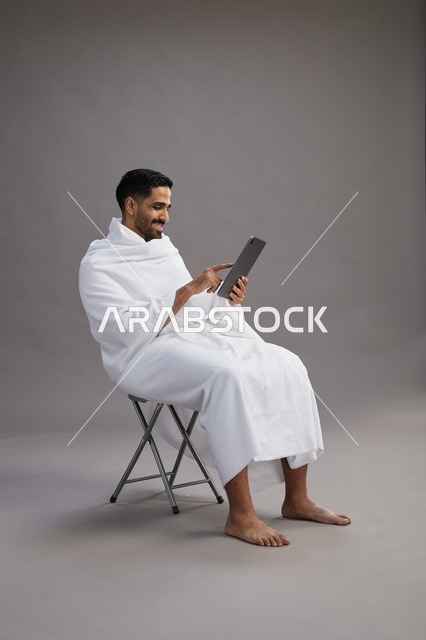 بورتريه لحاج سعودي بملابس الإحرام يحمل تابلت ويجلس على كرسي, الاستعداد لاداء مناسك الحج والعمره, خلفيه رماديه