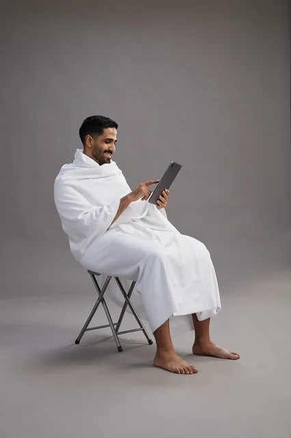 بورتريه لحاج سعودي بملابس الإحرام يحمل تابلت ويجلس على كرسي, الاستعداد لاداء مناسك الحج والعمره, خلفيه رماديه