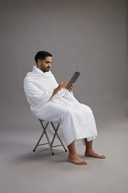 A portrait of a Saudi pilgrim in Ihram clothing holding a tablet and sitting on a chair, preparing to perform the rituals of Hajj and Umrah, with a gray background.