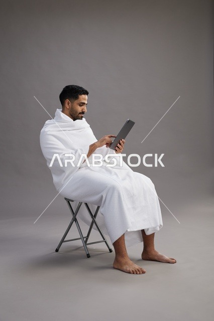 A portrait of a Saudi pilgrim in Ihram clothing holding a tablet and sitting on a chair, preparing to perform the rituals of Hajj and Umrah, with a gray background.