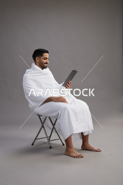 بورتريه لحاج سعودي بملابس الإحرام يحمل تابلت ويجلس على كرسي, الاستعداد لاداء مناسك الحج والعمره, خلفيه رماديه