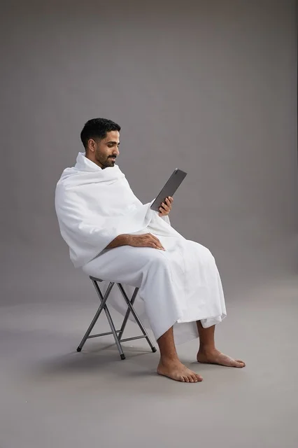 A portrait of a Saudi pilgrim in Ihram clothing holding a tablet and sitting on a chair, preparing to perform the rituals of Hajj and Umrah, with a gray background.