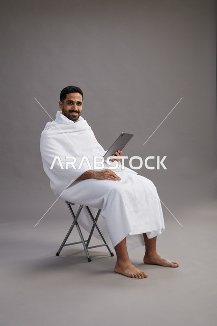 بورتريه لحاج سعودي بملابس الإحرام يحمل تابلت ويجلس على كرسي, الاستعداد لاداء مناسك الحج والعمره, خلفيه رماديه