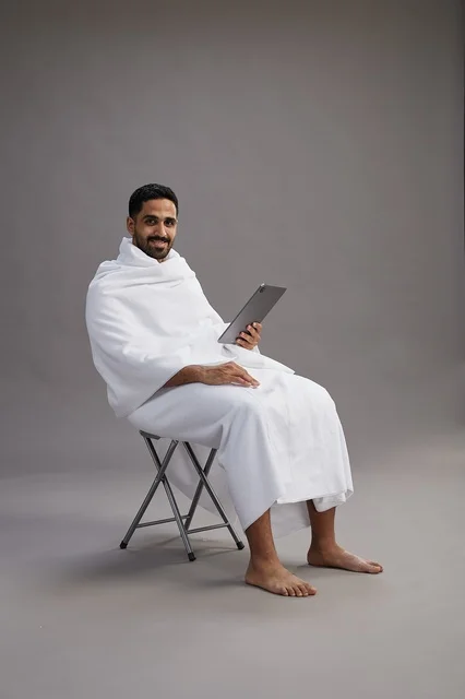 A portrait of a Saudi pilgrim in Ihram clothing holding a tablet and sitting on a chair, preparing to perform the rituals of Hajj and Umrah, with a gray background.