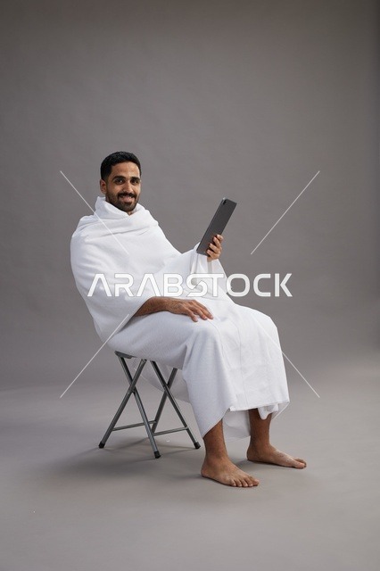 A portrait of a Saudi pilgrim in Ihram clothing holding a tablet and sitting on a chair, preparing to perform the rituals of Hajj and Umrah, with a gray background.