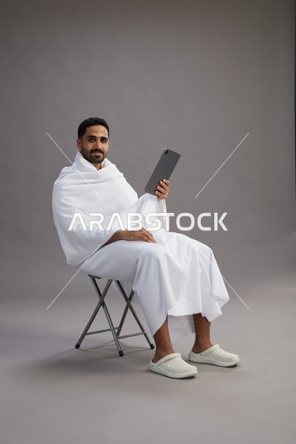 بورتريه لحاج سعودي بملابس الإحرام يحمل تابلت ويجلس على كرسي, الاستعداد لاداء مناسك الحج والعمره, خلفيه رماديه