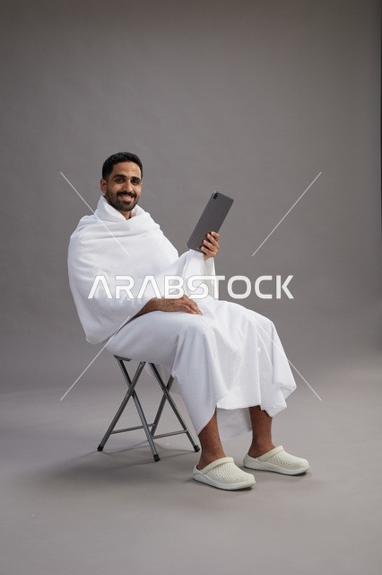 بورتريه لحاج سعودي بملابس الإحرام يحمل تابلت ويجلس على كرسي, الاستعداد لاداء مناسك الحج والعمره, خلفيه رماديه