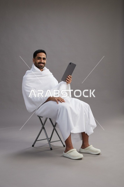 A portrait of a Saudi pilgrim in Ihram clothing holding a tablet and sitting on a chair, preparing to perform the rituals of Hajj and Umrah, with a gray background.