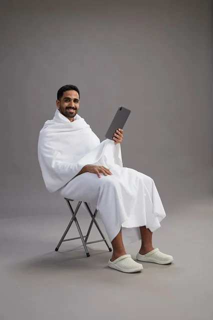 A portrait of a Saudi pilgrim in Ihram clothing holding a tablet and sitting on a chair, preparing to perform the rituals of Hajj and Umrah, with a gray background.
