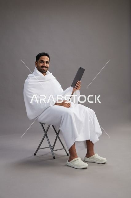 بورتريه لحاج سعودي بملابس الإحرام يحمل تابلت ويجلس على كرسي, الاستعداد لاداء مناسك الحج والعمره, خلفيه رماديه