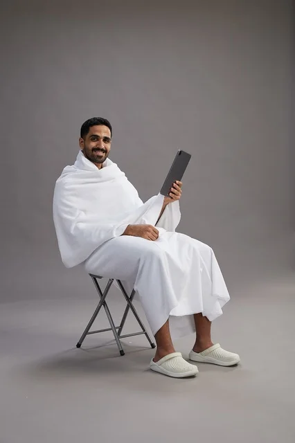 A portrait of a Saudi pilgrim in Ihram clothing holding a tablet and sitting on a chair, preparing to perform the rituals of Hajj and Umrah, with a gray background.