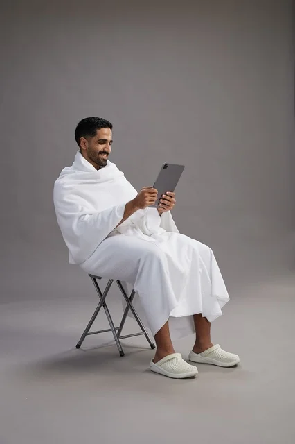 A portrait of a Saudi pilgrim in Ihram clothing holding a tablet and sitting on a chair, preparing to perform the rituals of Hajj and Umrah, with a gray background.