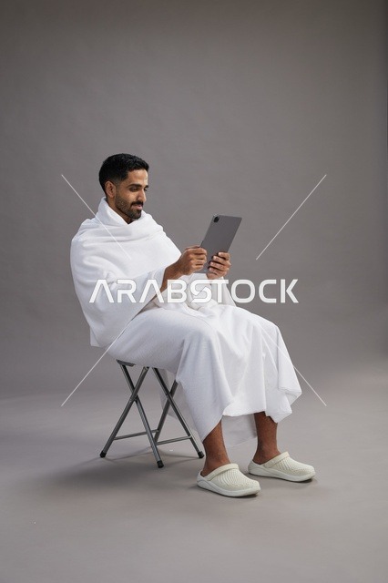 بورتريه لحاج سعودي بملابس الإحرام يحمل تابلت ويجلس على كرسي, الاستعداد لاداء مناسك الحج والعمره, خلفيه رماديه