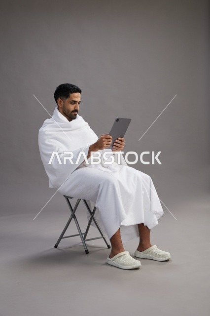 A portrait of a Saudi pilgrim in Ihram clothing holding a tablet and sitting on a chair, preparing to perform the rituals of Hajj and Umrah, with a gray background.