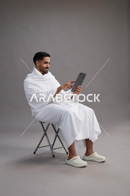 A portrait of a Saudi pilgrim in Ihram clothing holding a tablet and sitting on a chair, preparing to perform the rituals of Hajj and Umrah, with a gray background.