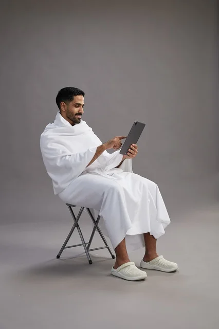 A portrait of a Saudi pilgrim in Ihram clothing holding a tablet and sitting on a chair, preparing to perform the rituals of Hajj and Umrah, with a gray background.