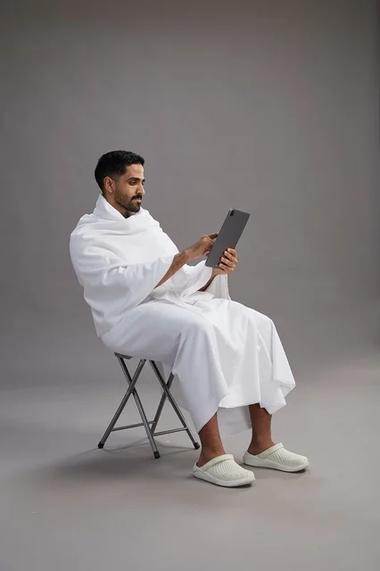 بورتريه لحاج سعودي بملابس الإحرام يحمل تابلت ويجلس على كرسي, الاستعداد لاداء مناسك الحج والعمره, خلفيه رماديه