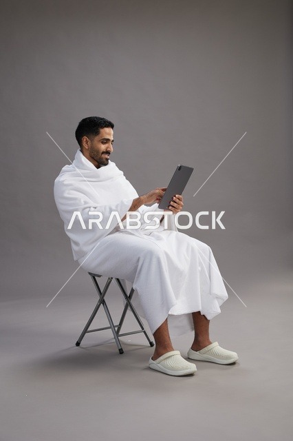 بورتريه لحاج سعودي بملابس الإحرام يحمل تابلت ويجلس على كرسي, الاستعداد لاداء مناسك الحج والعمره, خلفيه رماديه