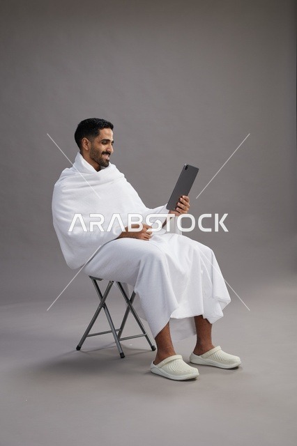 بورتريه لحاج سعودي بملابس الإحرام يحمل تابلت ويجلس على كرسي, الاستعداد لاداء مناسك الحج والعمره, خلفيه رماديه