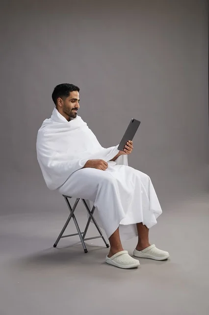 A portrait of a Saudi pilgrim in Ihram clothing holding a tablet and sitting on a chair, preparing to perform the rituals of Hajj and Umrah, with a gray background.