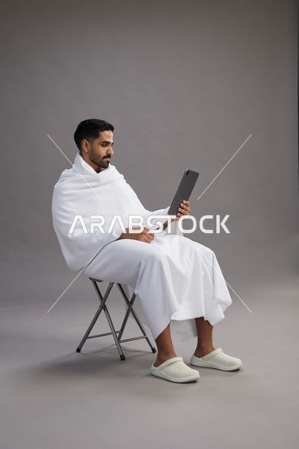 بورتريه لحاج سعودي بملابس الإحرام يحمل تابلت ويجلس على كرسي, الاستعداد لاداء مناسك الحج والعمره, خلفيه رماديه