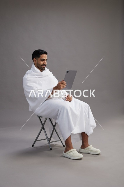 A portrait of a Saudi pilgrim in Ihram clothing holding a tablet and sitting on a chair, preparing to perform the rituals of Hajj and Umrah, with a gray background.