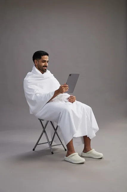 بورتريه لحاج سعودي بملابس الإحرام يحمل تابلت ويجلس على كرسي, الاستعداد لاداء مناسك الحج والعمره, خلفيه رماديه