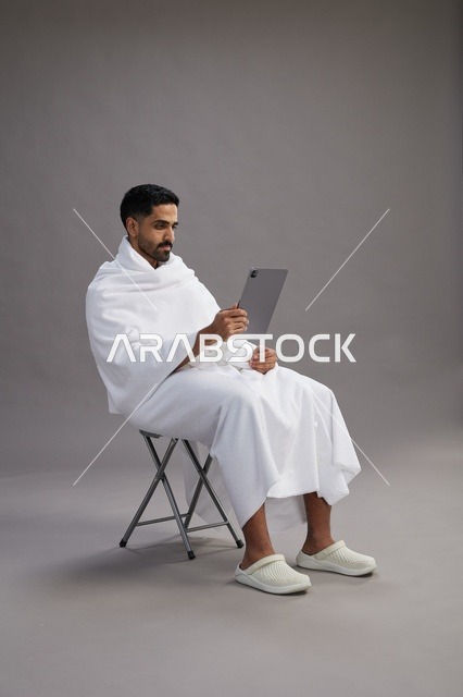 بورتريه لحاج سعودي بملابس الإحرام يحمل تابلت ويجلس على كرسي, الاستعداد لاداء مناسك الحج والعمره, خلفيه رماديه