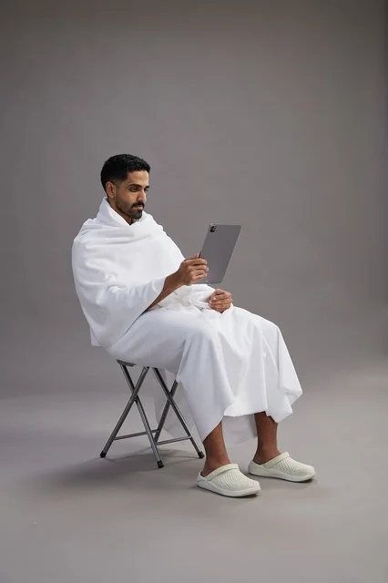 بورتريه لحاج سعودي بملابس الإحرام يحمل تابلت ويجلس على كرسي, الاستعداد لاداء مناسك الحج والعمره, خلفيه رماديه