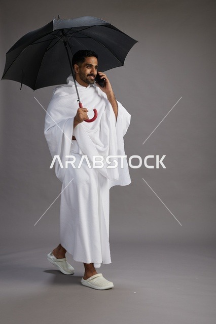 بورتريه لحاج  عربي سعودي  خليجي بملابس الإحرام يحمل مظله, الذهاب لاداء مناسك الحج والعمره, استخدام الهاتف المحمول  كامل الجسم, خلفيه رماديه
