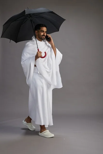 A portrait of an Arab Saudi Gulf pilgrim in Ihram clothing holding an umbrella, going to perform the rituals of Hajj and Umrah, using a mobile phone, full body, gray background.