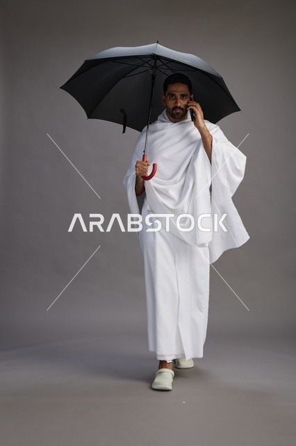 بورتريه لحاج  عربي سعودي  خليجي بملابس الإحرام يحمل مظله, الذهاب لاداء مناسك الحج والعمره, استخدام الهاتف المحمول  كامل الجسم, خلفيه رماديه