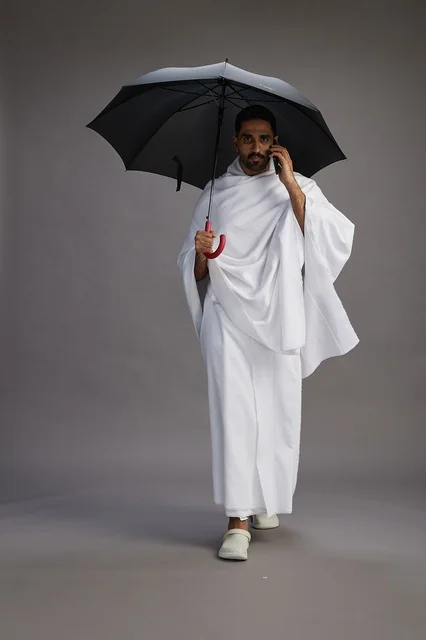 A portrait of an Arab Saudi Gulf pilgrim in Ihram clothing holding an umbrella, going to perform the rituals of Hajj and Umrah, using a mobile phone, full body, gray background.