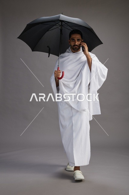 بورتريه لحاج  عربي سعودي  خليجي بملابس الإحرام يحمل مظله, الذهاب لاداء مناسك الحج والعمره, استخدام الهاتف المحمول  كامل الجسم, خلفيه رماديه