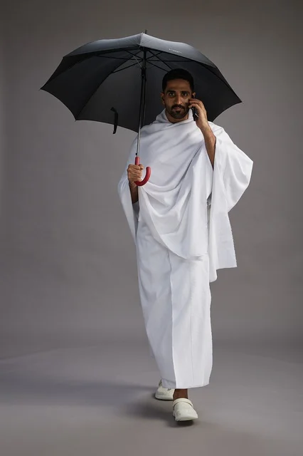 A portrait of an Arab Saudi Gulf pilgrim in Ihram clothing holding an umbrella, going to perform the rituals of Hajj and Umrah, using a mobile phone, full body, gray background.