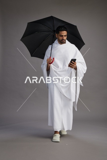 بورتريه لحاج  عربي سعودي  خليجي بملابس الإحرام يحمل مظله, الذهاب لاداء مناسك الحج والعمره, استخدام الهاتف المحمول  كامل الجسم, خلفيه رماديه