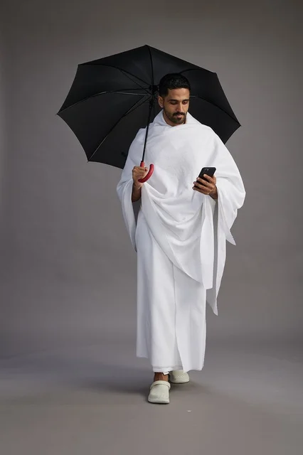 A portrait of an Arab Saudi Gulf pilgrim in Ihram clothing holding an umbrella, going to perform the rituals of Hajj and Umrah, using a mobile phone, full body, gray background.