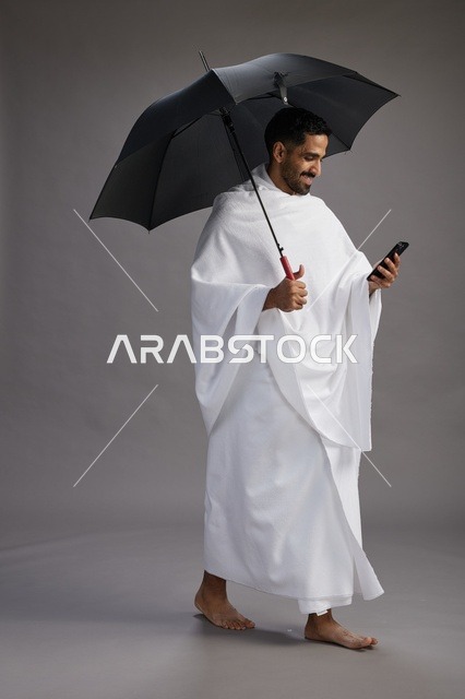 بورتريه لحاج  عربي سعودي  خليجي بملابس الإحرام يحمل مظله, الذهاب لاداء مناسك الحج والعمره, استخدام الهاتف المحمول  كامل الجسم, خلفيه رماديه