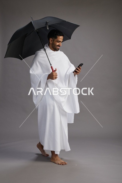 بورتريه لحاج  عربي سعودي  خليجي بملابس الإحرام يحمل مظله, الذهاب لاداء مناسك الحج والعمره, استخدام الهاتف المحمول  كامل الجسم, خلفيه رماديه