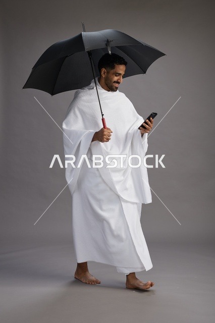 بورتريه لحاج  عربي سعودي  خليجي بملابس الإحرام يحمل مظله, الذهاب لاداء مناسك الحج والعمره, استخدام الهاتف المحمول  كامل الجسم, خلفيه رماديه